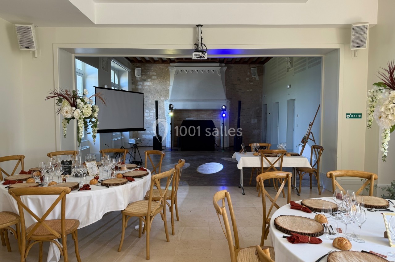 Salle de réception lumineuse avec tables dressées, écran de projection, projecteurs et décoration florale.