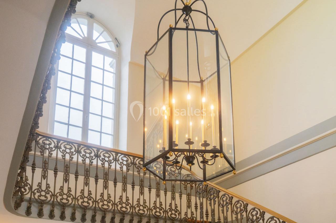 Lustre suspendu en verre et métal éclairé, au-dessus d'un escalier avec une rampe en fer forgé, près d'une grande fenêtre.