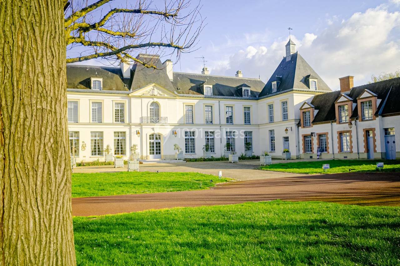 Façade d'un château blanc avec toit en ardoise, entouré d'une pelouse et d'une allée pavée, sous un ciel dégagé.