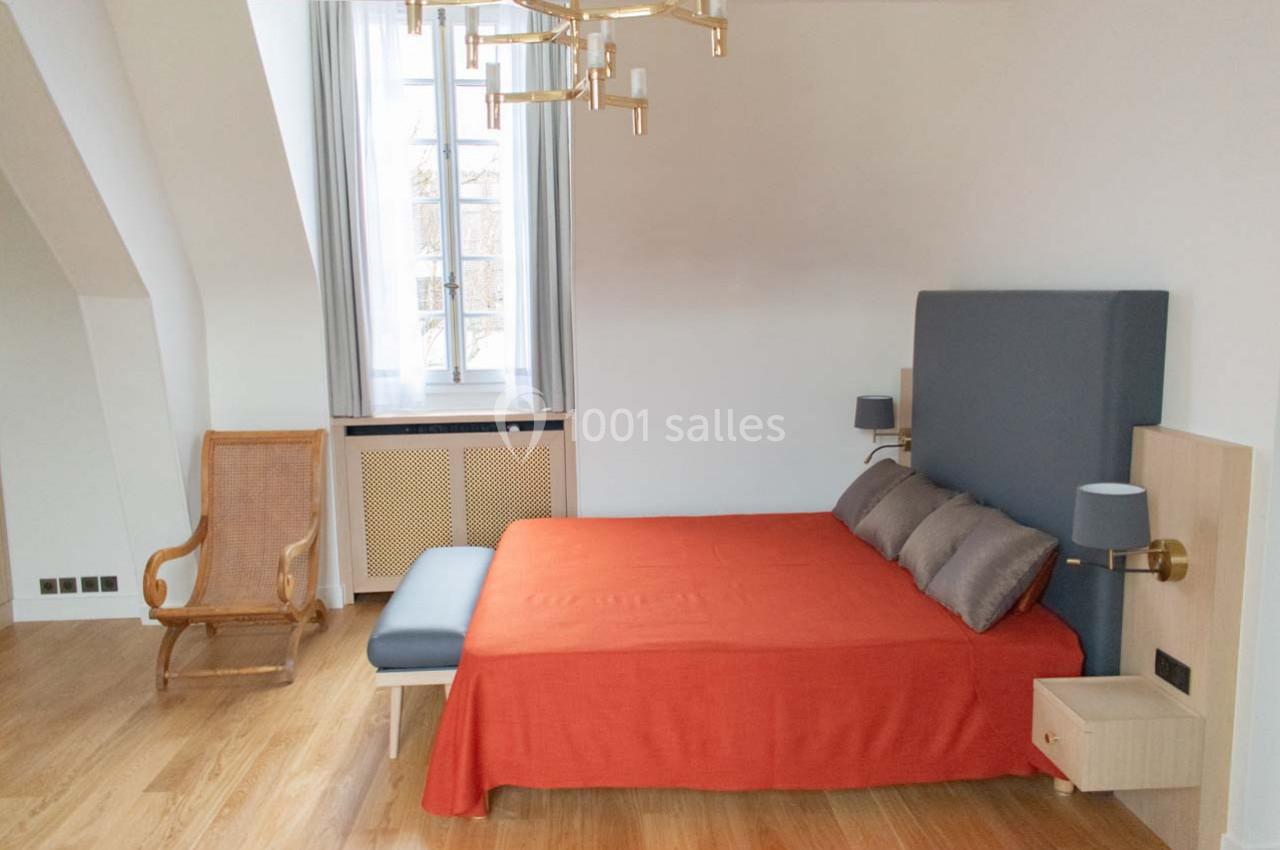 Chambre lumineuse avec lit double, couverture orange, fauteuil en bois, parquet et fenêtre encadrée de rideaux blancs.
