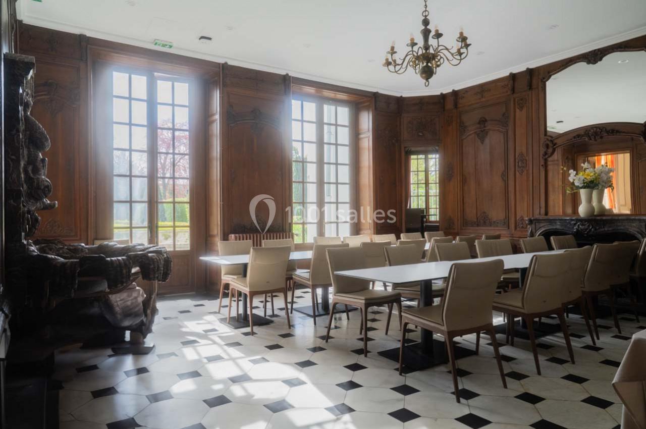 Salle lumineuse avec des murs en bois sculpté, des fenêtres hautes, des tables blanches et des chaises beiges.