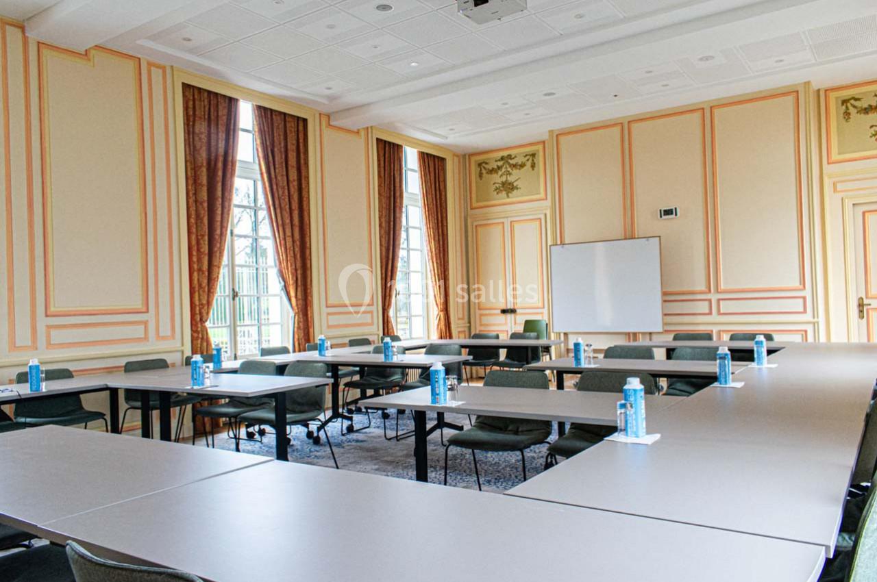 Salle de réunion lumineuse avec tables disposées en U, chaises, tableau blanc et bouteilles d'eau sur les tables.