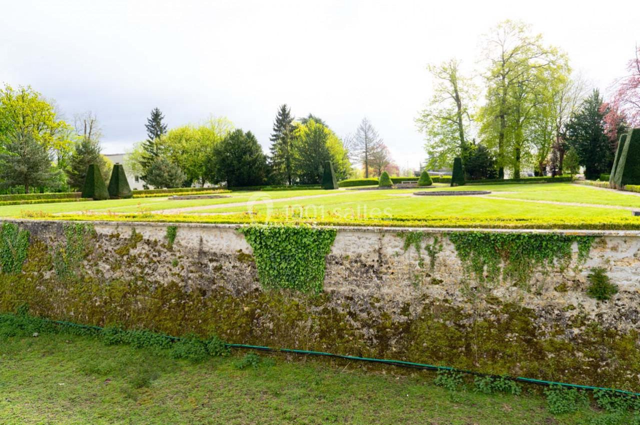 Mur en pierre recouvert de mousse et de lierre, surmonté d'un jardin avec pelouse et arbres en arrière-plan.