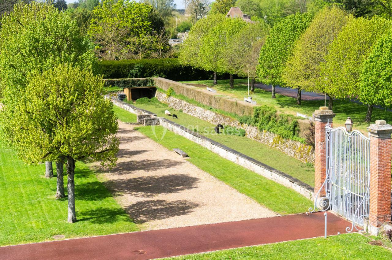 Allée bordée d'arbres et de pelouses, avec un mur en pierre et une grille métallique à droite, par temps ensoleillé.