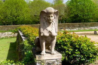 Statue de sphinx en pierre posée sur un socle, entourée de verdure dans un jardin.