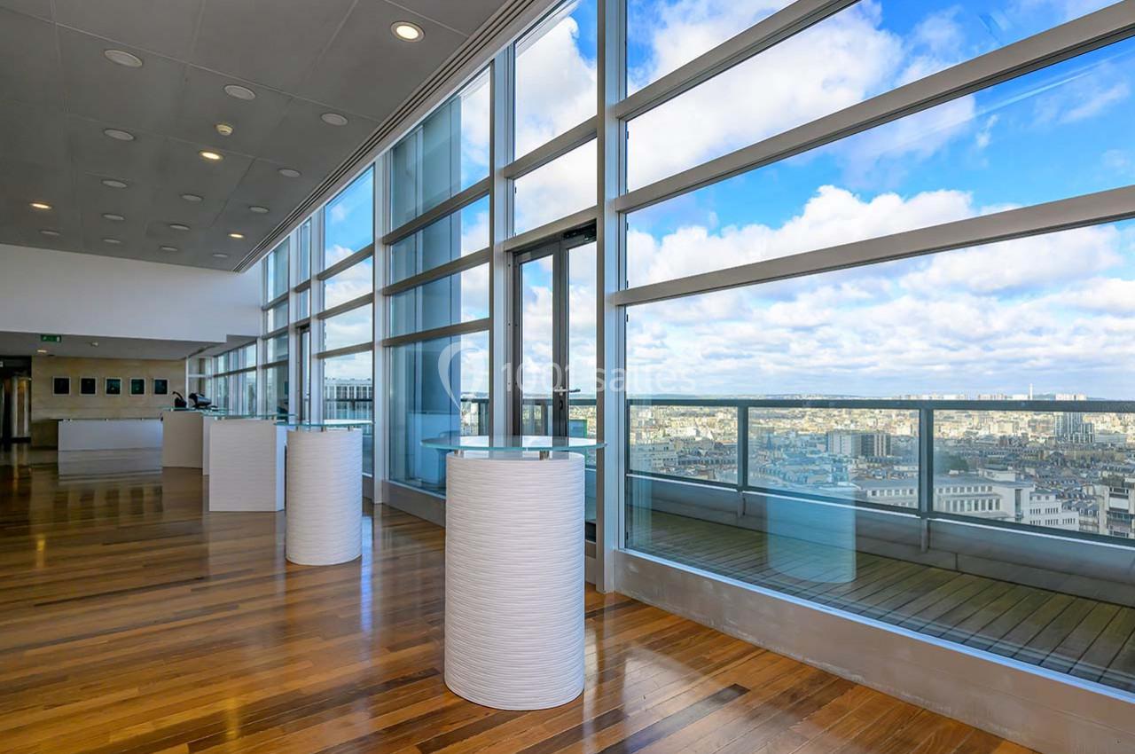 Salle lumineuse avec de grandes baies vitrées offrant une vue panoramique sur une ville sous un ciel partiellement nuageux.