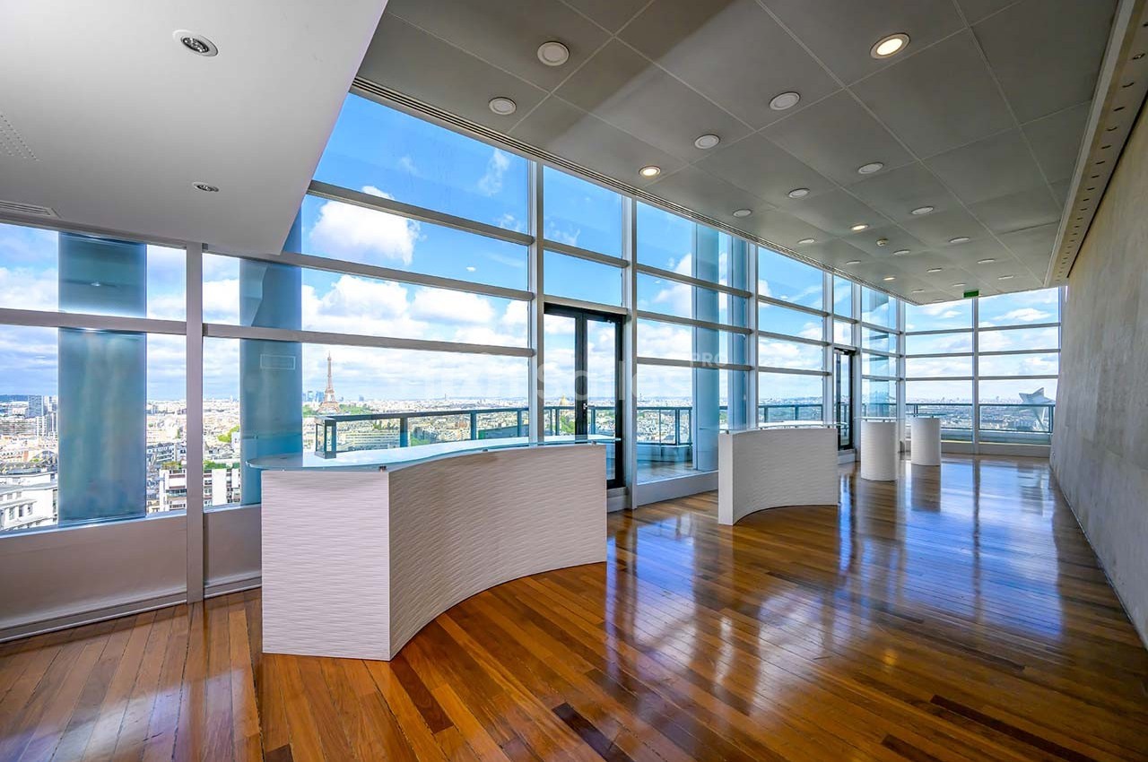 Salle lumineuse avec de grandes baies vitrées offrant une vue panoramique sur une ville, parquet en bois au sol.