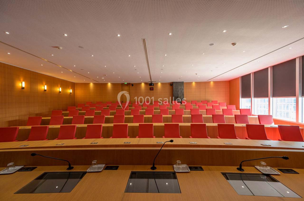 Salle de conférence vide avec rangées de sièges rouges, pupitres équipés de micros et éclairage tamisé.