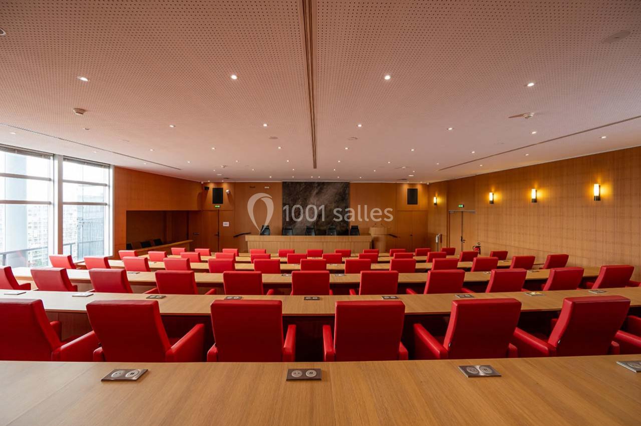 Salle de conférence moderne avec sièges rouges, tables en bois et éclairage intégré, vue vers l'estrade centrale.