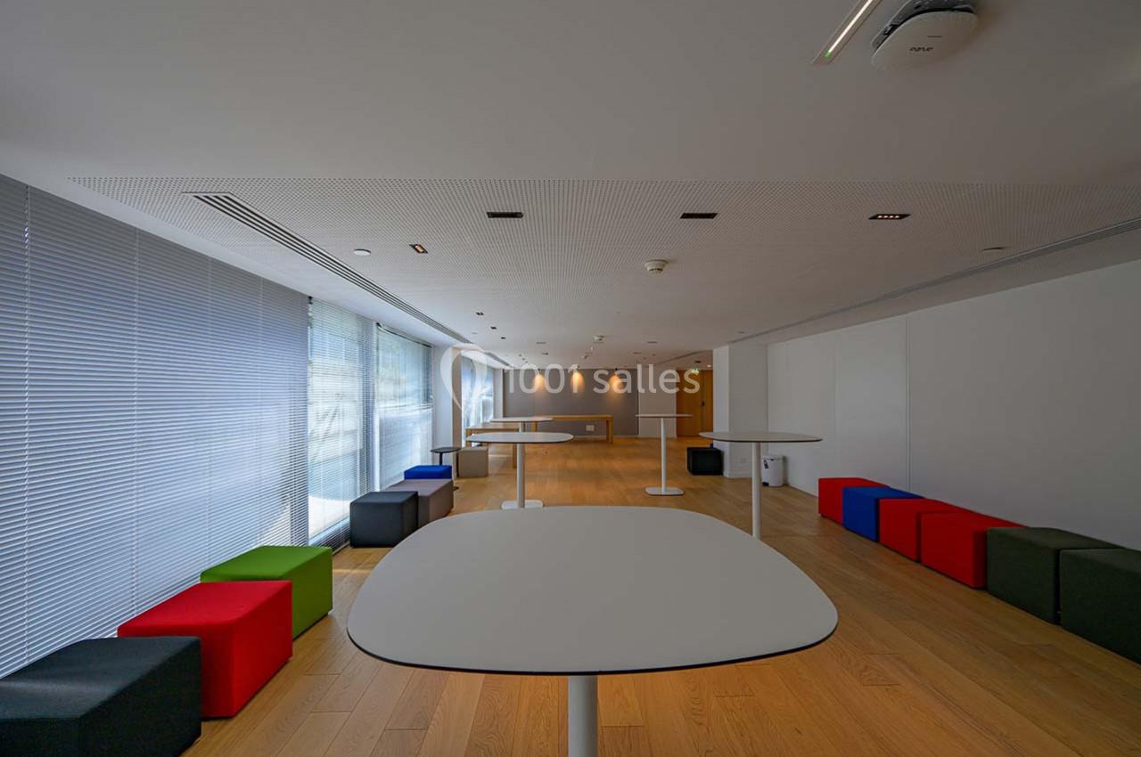 Salle lumineuse avec tables blanches, tabourets colorés et parquet en bois clair, bordée de grandes fenêtres.