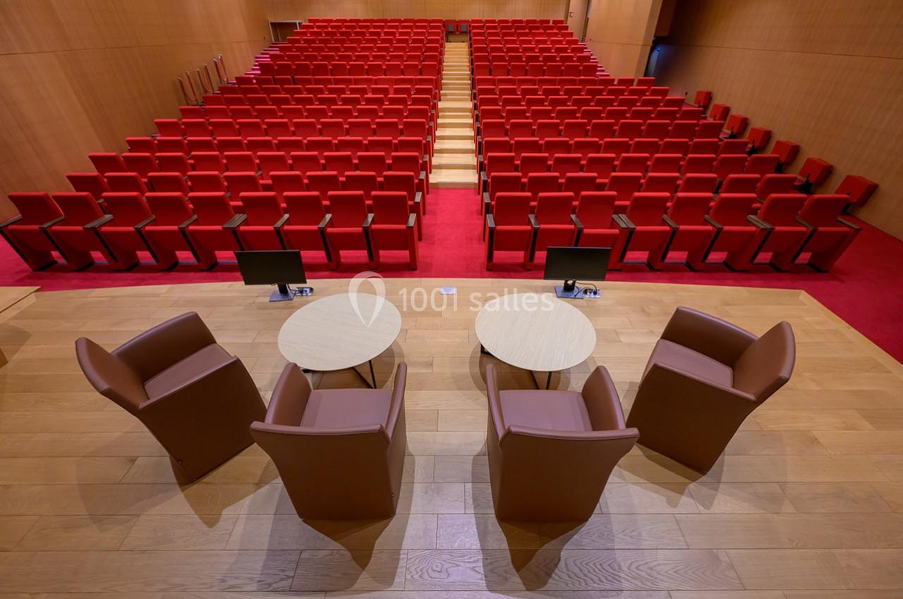 Salle de conférence avec rangées de sièges rouges, vue depuis une scène équipée de fauteuils et tables rondes.