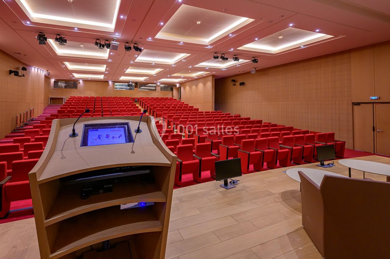 Vue depuis un pupitre sur un amphithéâtre vide avec des sièges rouges et un éclairage au plafond.