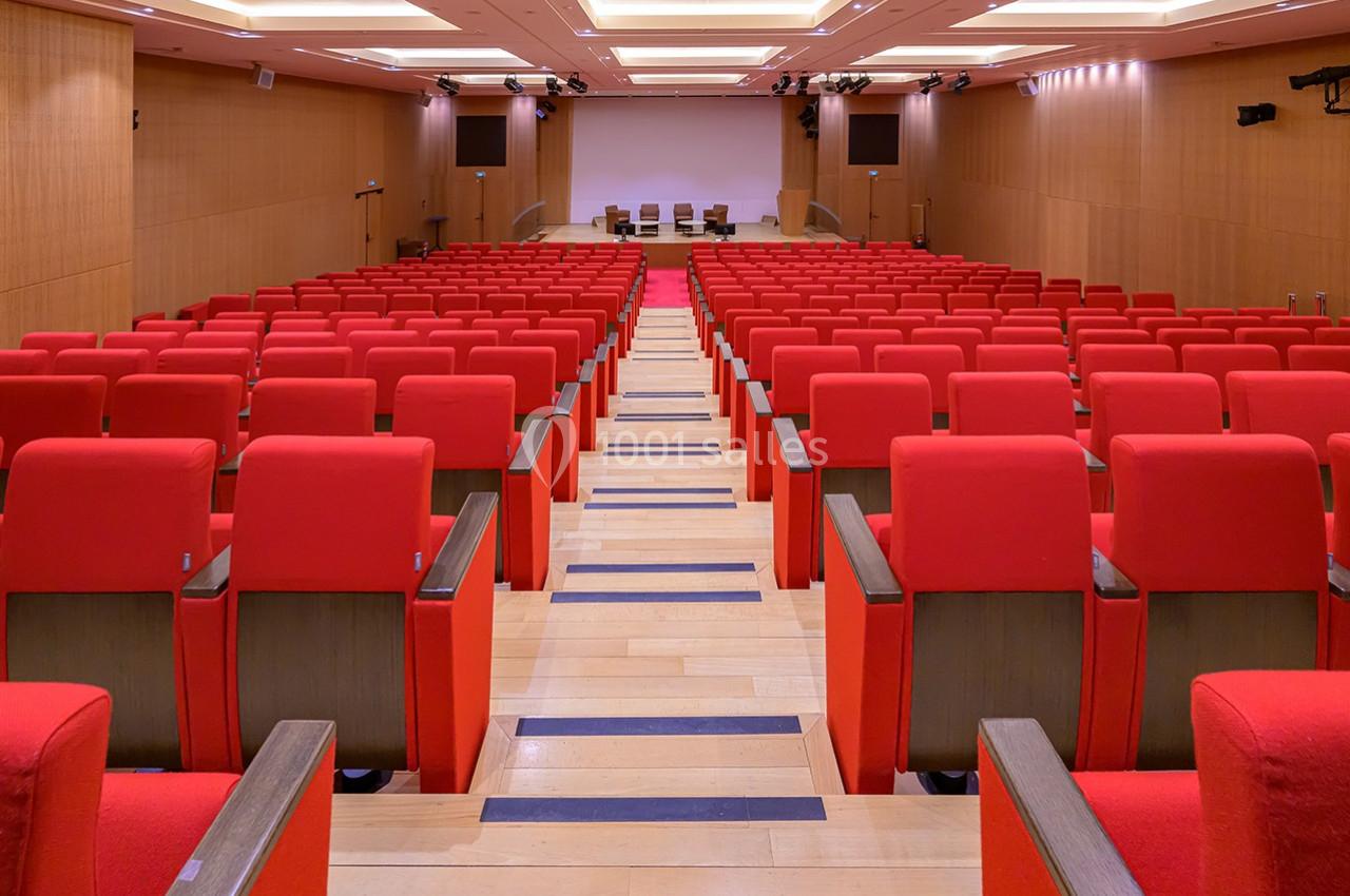 Salle de conférence avec des rangées de sièges rouges alignés face à un écran et une estrade au fond.