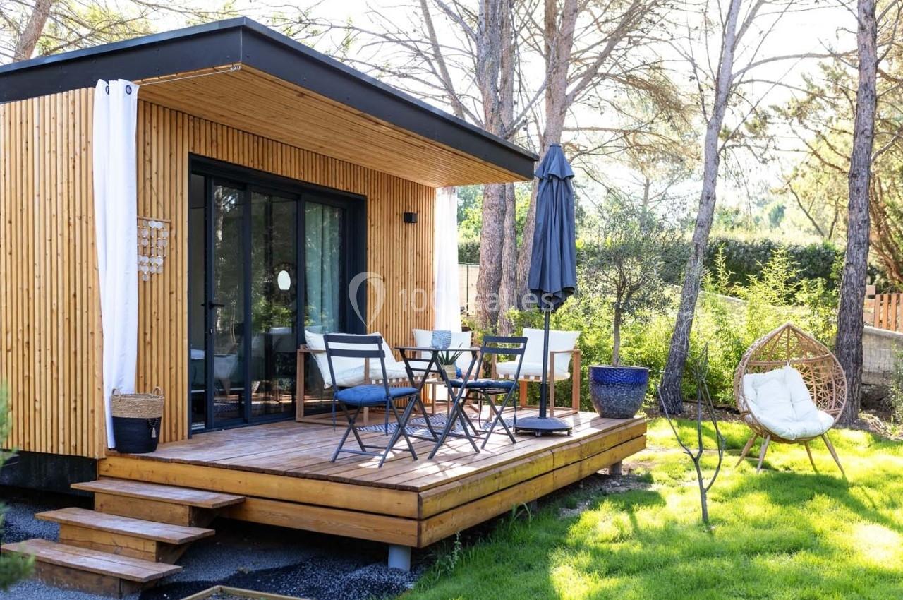 Terrasse en bois avec mobilier de jardin devant une petite maison moderne entourée de verdure et d'arbres.