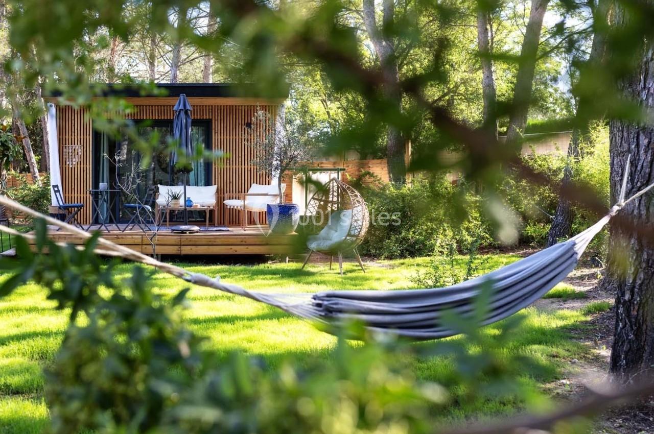 Terrasse en bois avec mobilier de jardin, entourée de verdure, vue à travers des branches et un hamac au premier plan.