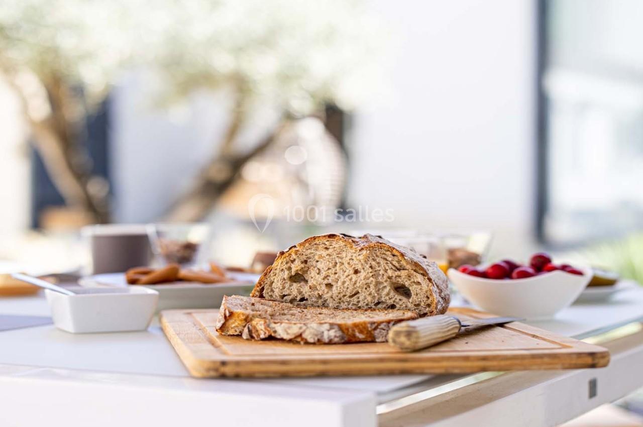 Tranche de pain rustique sur une planche en bois, entourée de bols de fruits et d'accompagnements sur une table extérieure.