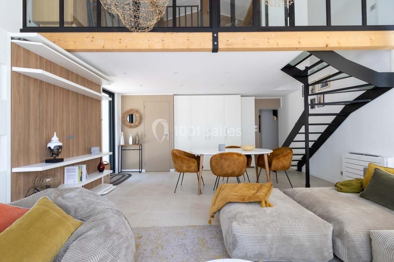 Salon moderne avec mezzanine, canapé beige, table à manger entourée de chaises moutarde et escalier en métal noir.