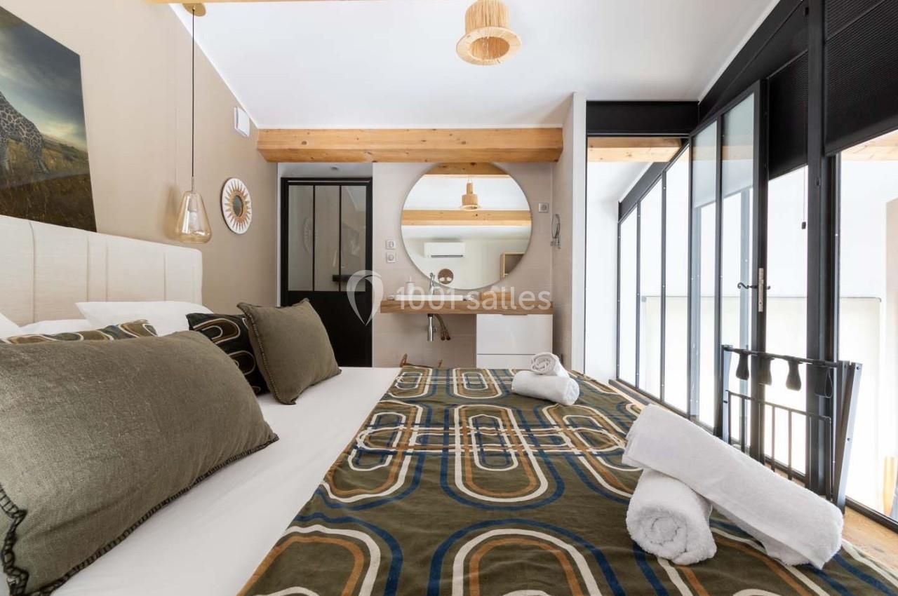 Chambre lumineuse avec lit double, linge de lit à motifs, miroir rond et salle de bain attenante visible en arrière-plan.