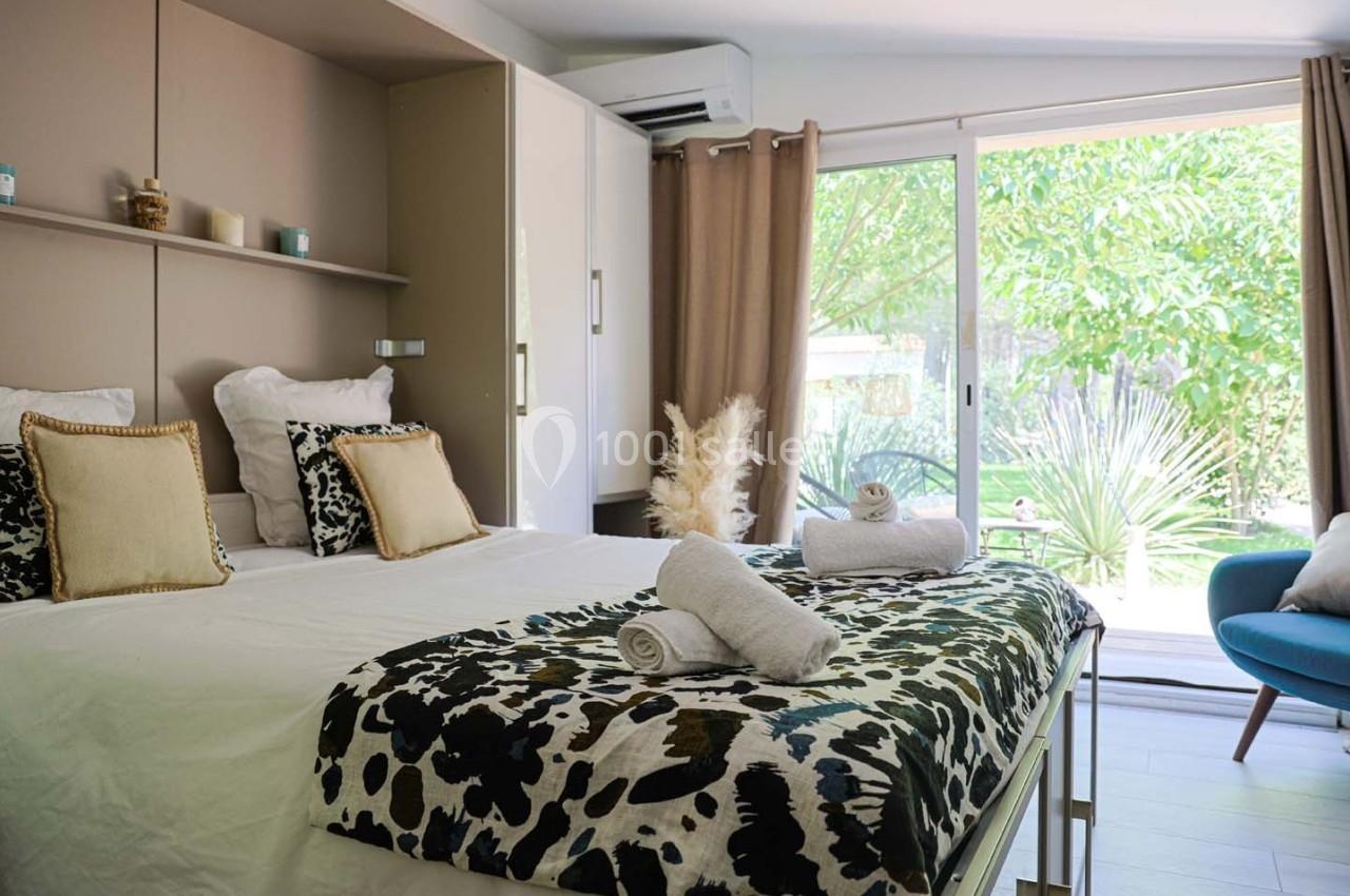 Chambre lumineuse avec lit double, coussins décoratifs, serviettes pliées et grande baie vitrée donnant sur un jardin.