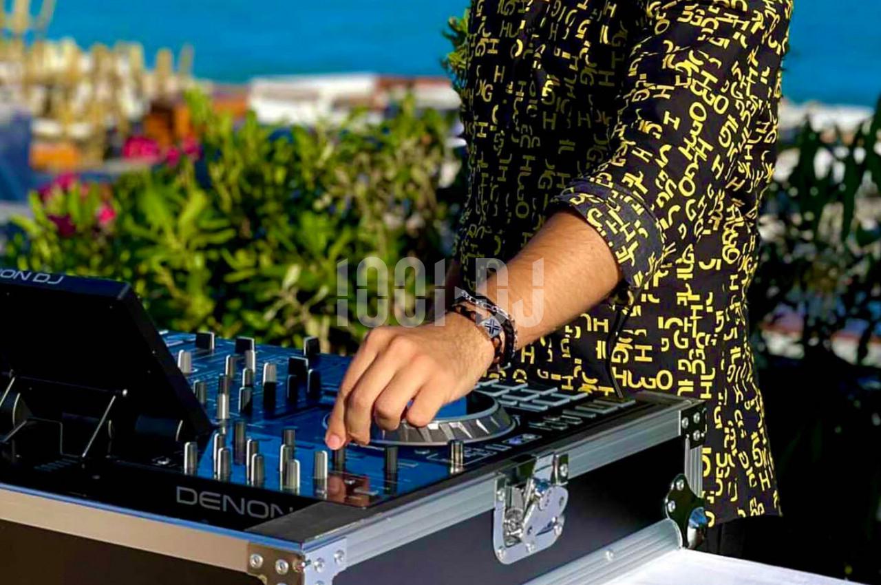 Un DJ ajuste une table de mixage en plein air, entouré de verdure et avec une vue sur la mer en arrière-plan.