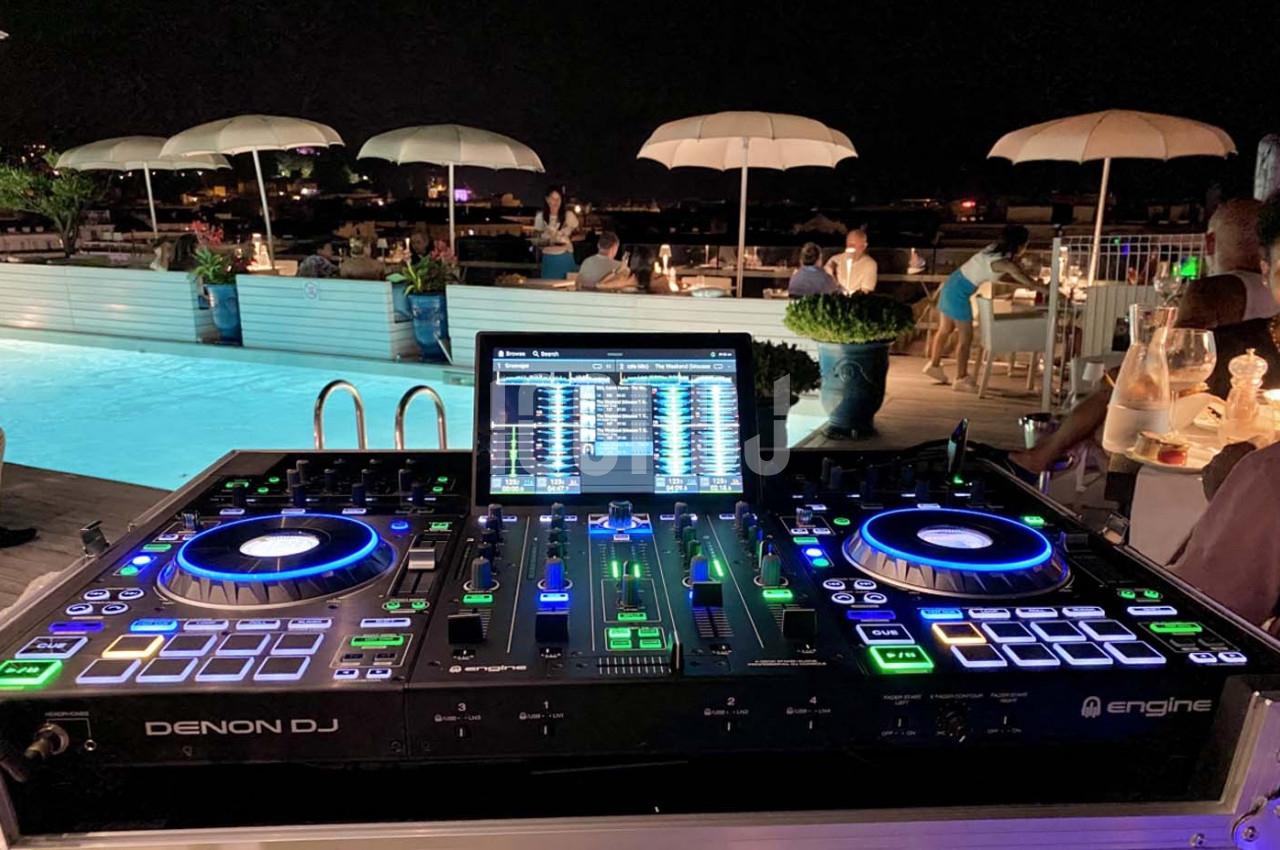 Table de mixage DJ éclairée près d'une piscine, avec des invités assis sous des parasols en arrière-plan.