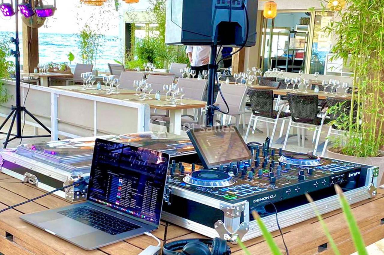 Table de mixage et ordinateur portable installés sur une terrasse en bord de mer, avec des tables dressées en arrière-plan.