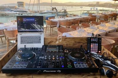 Poste de DJ avec platines, ordinateur et contrôleur, installé en terrasse face à un port de plaisance au coucher du soleil.