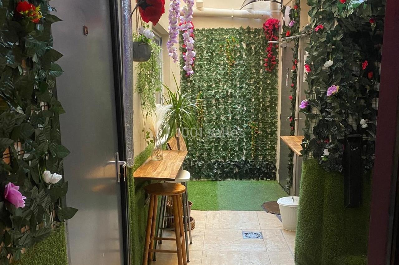Couloir étroit décoré de plantes artificielles, fleurs colorées et comptoirs en bois avec tabourets.