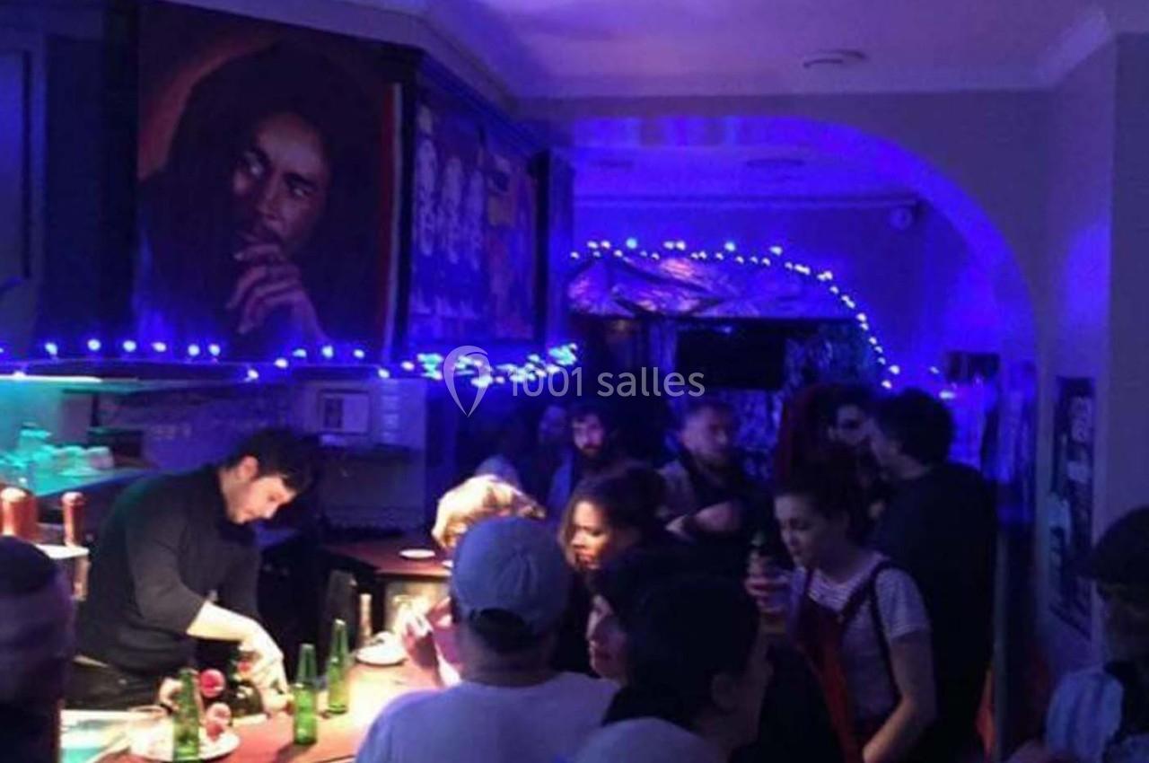 Un bar animé avec des clients discutant et un barman servant des boissons, éclairé par des lumières bleues.