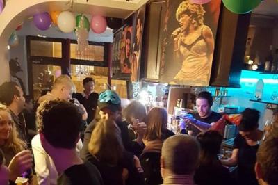 Façade d'un café avec une table extérieure en bois, revêtement en gazon synthétique et intérieur éclairé visible.