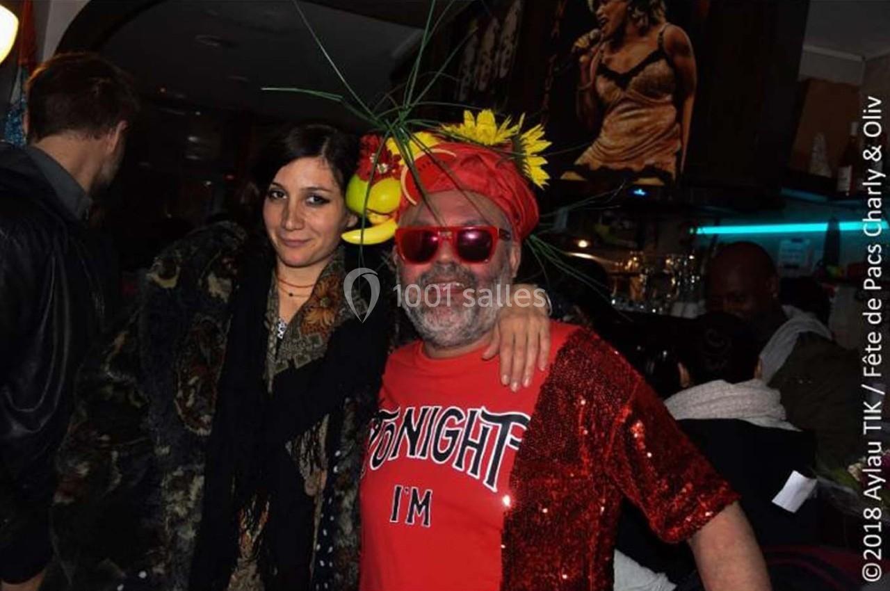 Un homme souriant en tenue festive avec un chapeau décoré et une femme posant à ses côtés dans un bar animé.