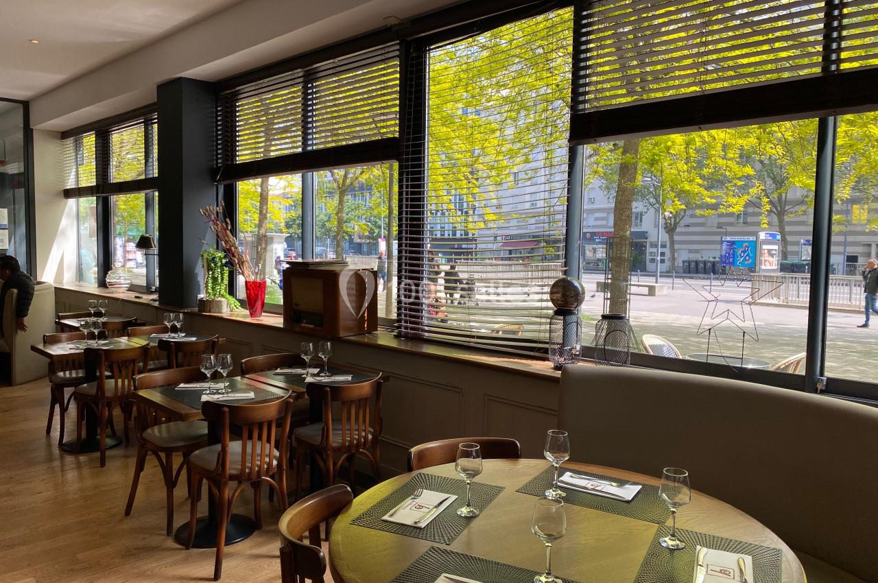 Salle de restaurant lumineuse avec des tables dressées, des chaises en bois et de grandes fenêtres donnant sur la rue.