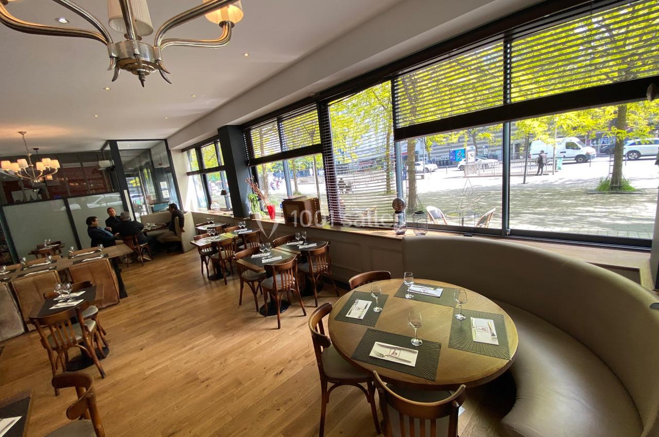 Salle de restaurant lumineuse avec grandes baies vitrées, tables en bois et banquettes arrondies près des fenêtres.