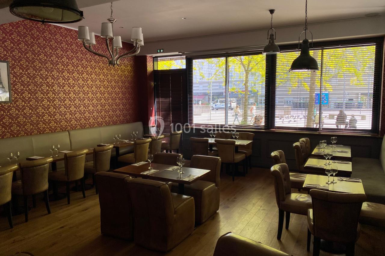 Salle de restaurant vide avec tables dressées, éclairée par la lumière naturelle entrant par de grandes fenêtres avec stores.