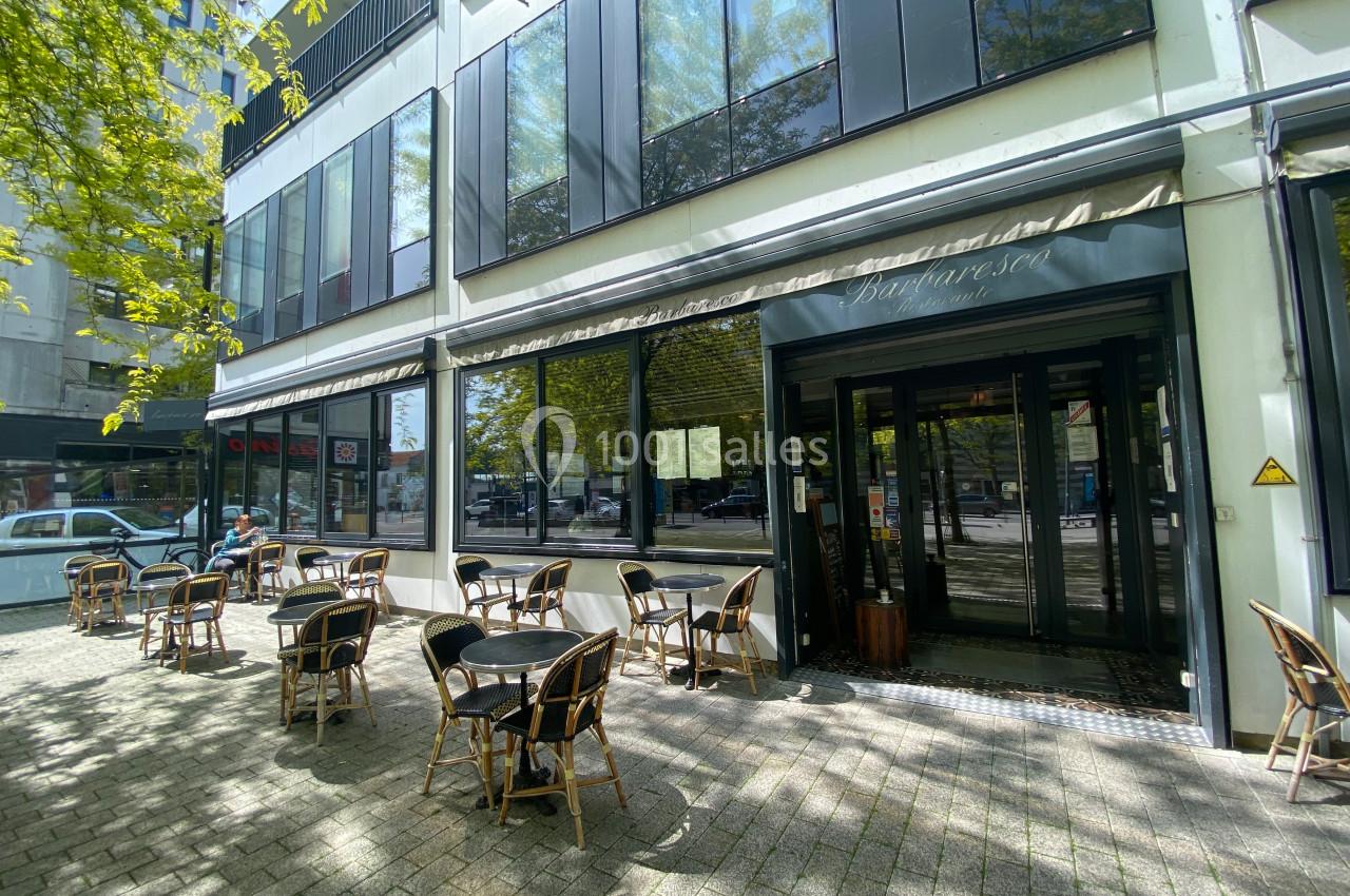 Terrasse d'un café avec des tables et chaises en extérieur, située devant une façade vitrée sous un auvent.