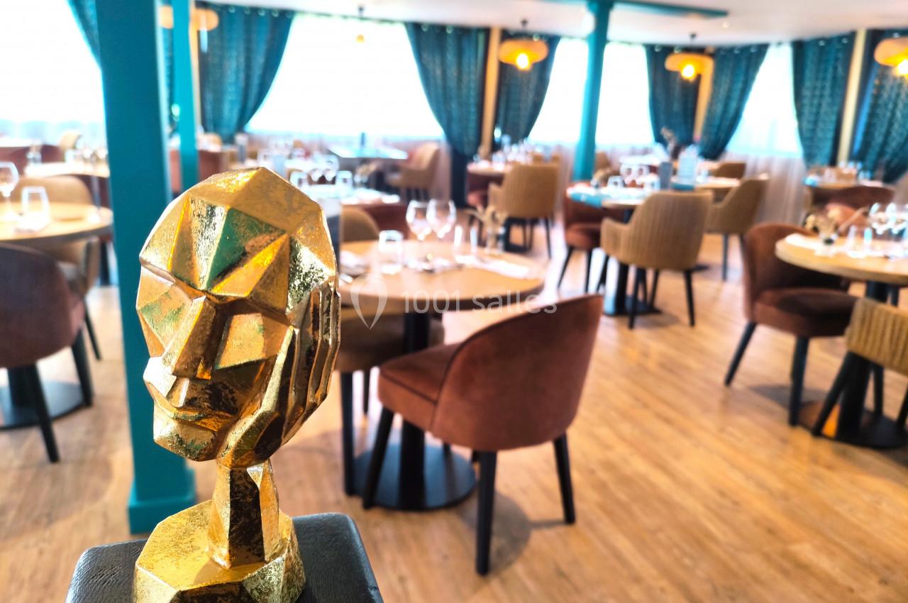Statue dorée géométrique en premier plan dans un restaurant élégant avec tables dressées et rideaux bleus.
