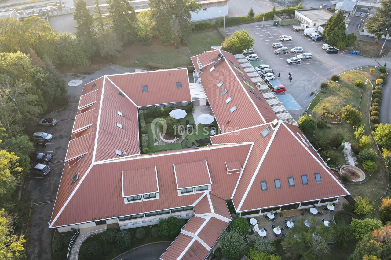 Vue aérienne d'un bâtiment en forme de U avec toit rouge, entouré de végétation, parking et espaces extérieurs aménagés.