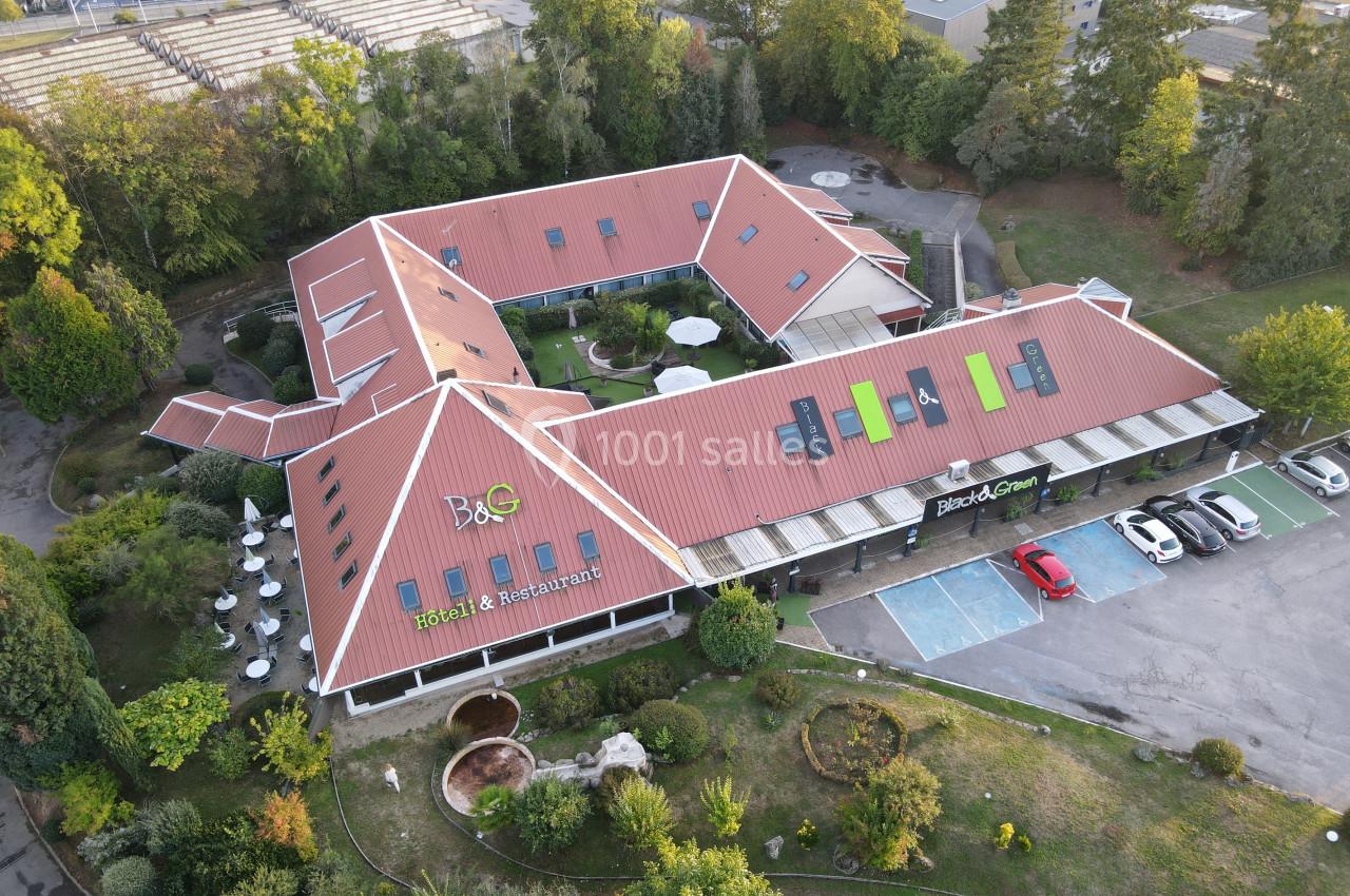 Vue aérienne d'un hôtel-restaurant avec toit rouge, terrasses extérieures et parking adjacent entouré de verdure.