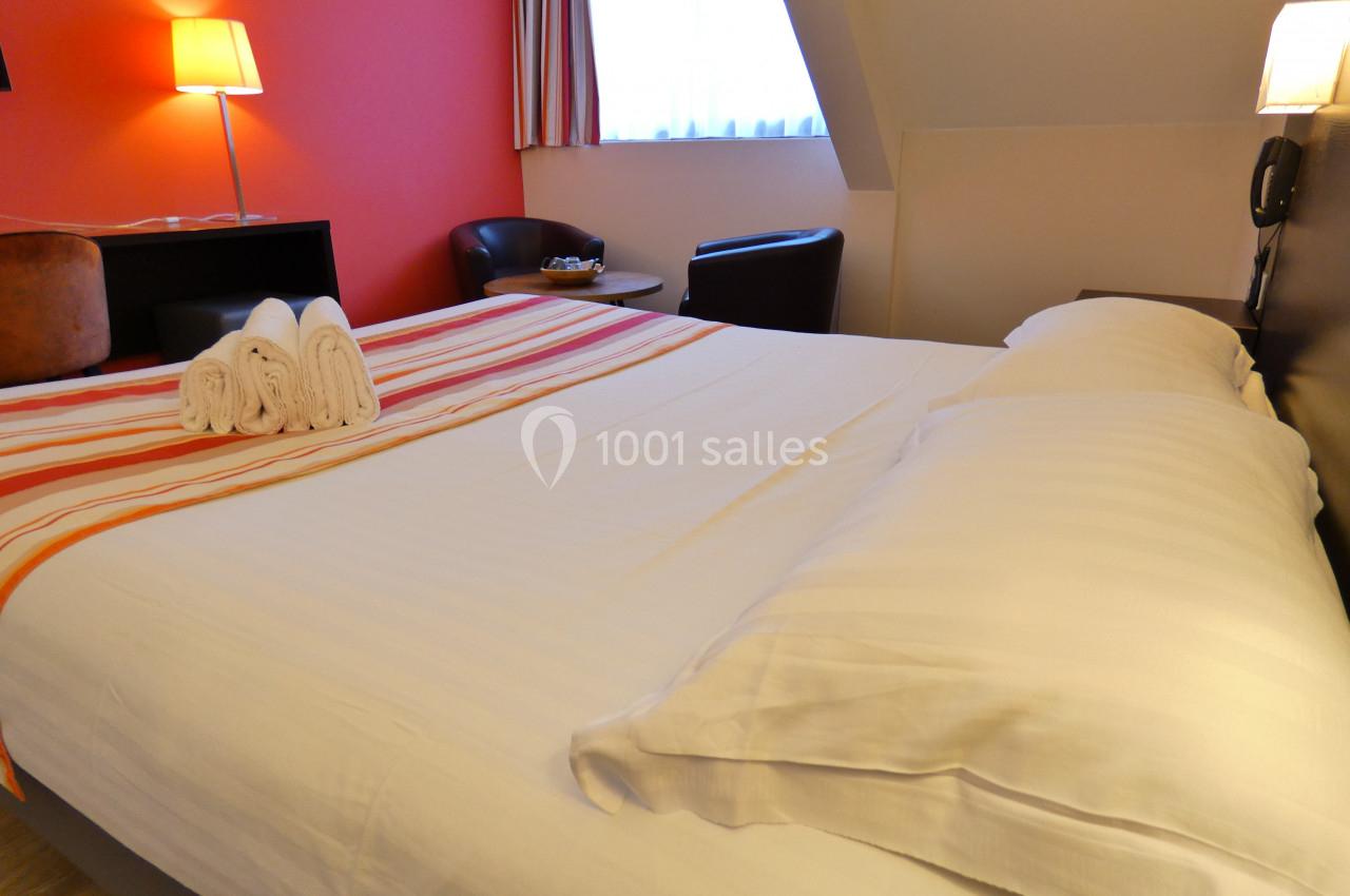 Lit double avec draps blancs, serviettes pliées, deux lampes allumées et un fauteuil près d'une fenêtre.