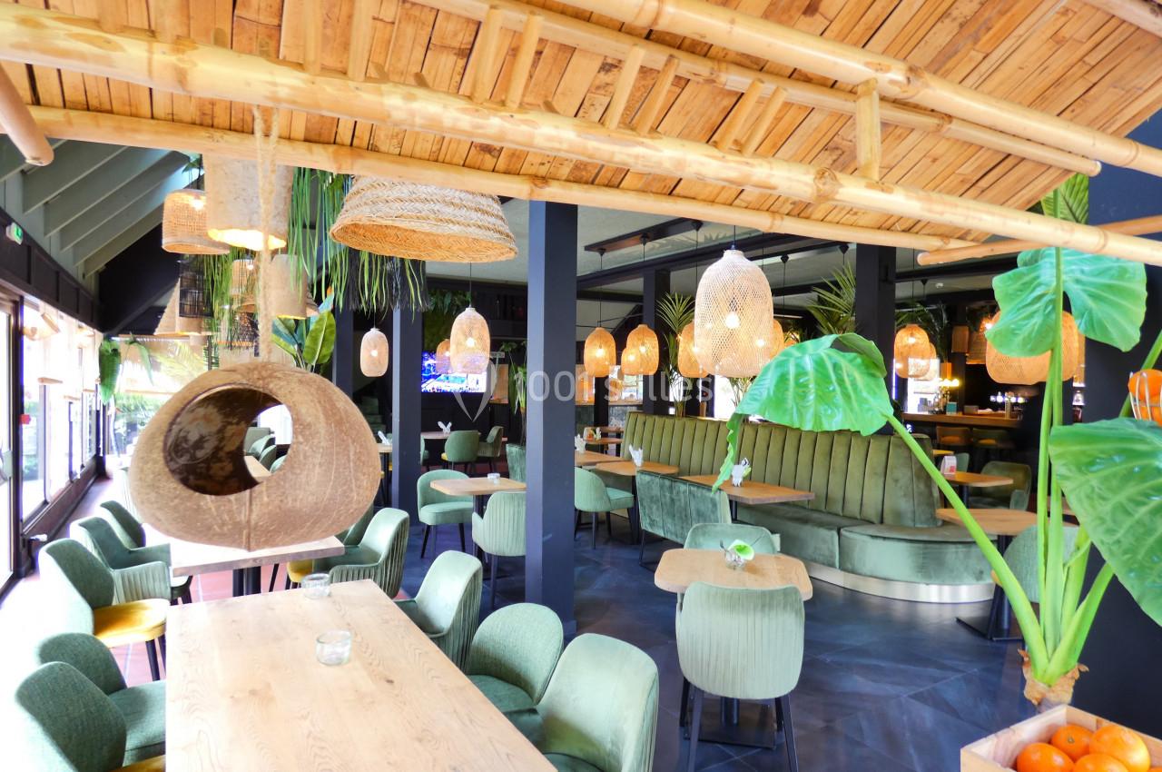 Intérieur d'un restaurant avec des tables en bois, des chaises vertes, des luminaires en osier et une décoration tropicale.