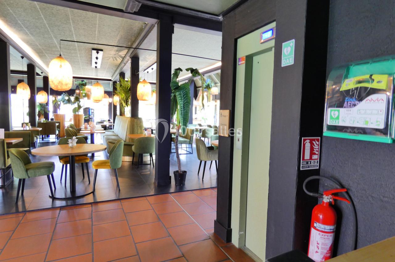 Salle de restaurant lumineuse avec des tables, des chaises vertes, un ascenseur et un extincteur au premier plan.