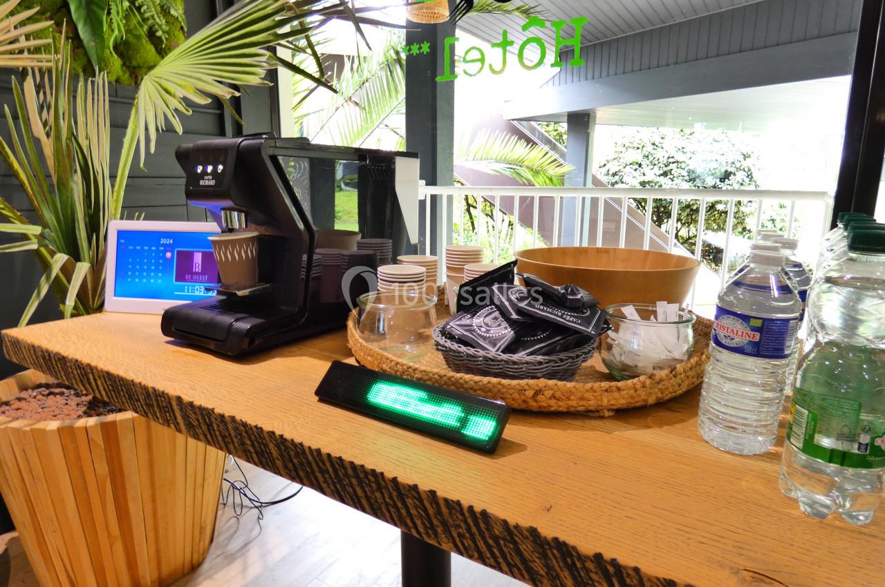 Machine à café avec gobelets, serviettes et bouteilles d'eau disposés sur une table en bois dans un espace lumineux.