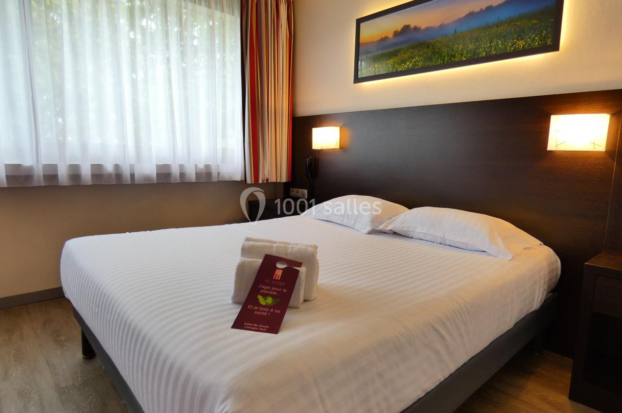 Lit double avec draps blancs dans une chambre lumineuse, décorée d'un tableau et éclairée par une lampe murale.