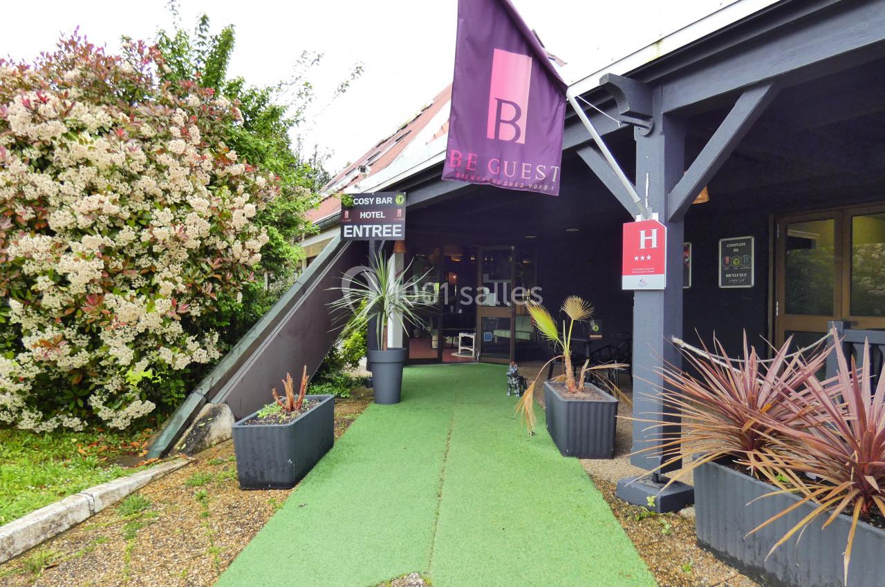 Entrée d'un hôtel avec un tapis vert, des plantes en pot et une bannière violette portant le logo ’Be Guest’.
