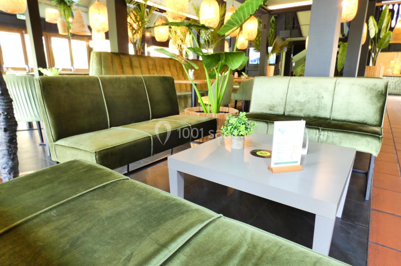 Canapés verts en velours autour d'une table basse grise dans un espace lumineux avec des plantes et des suspensions.