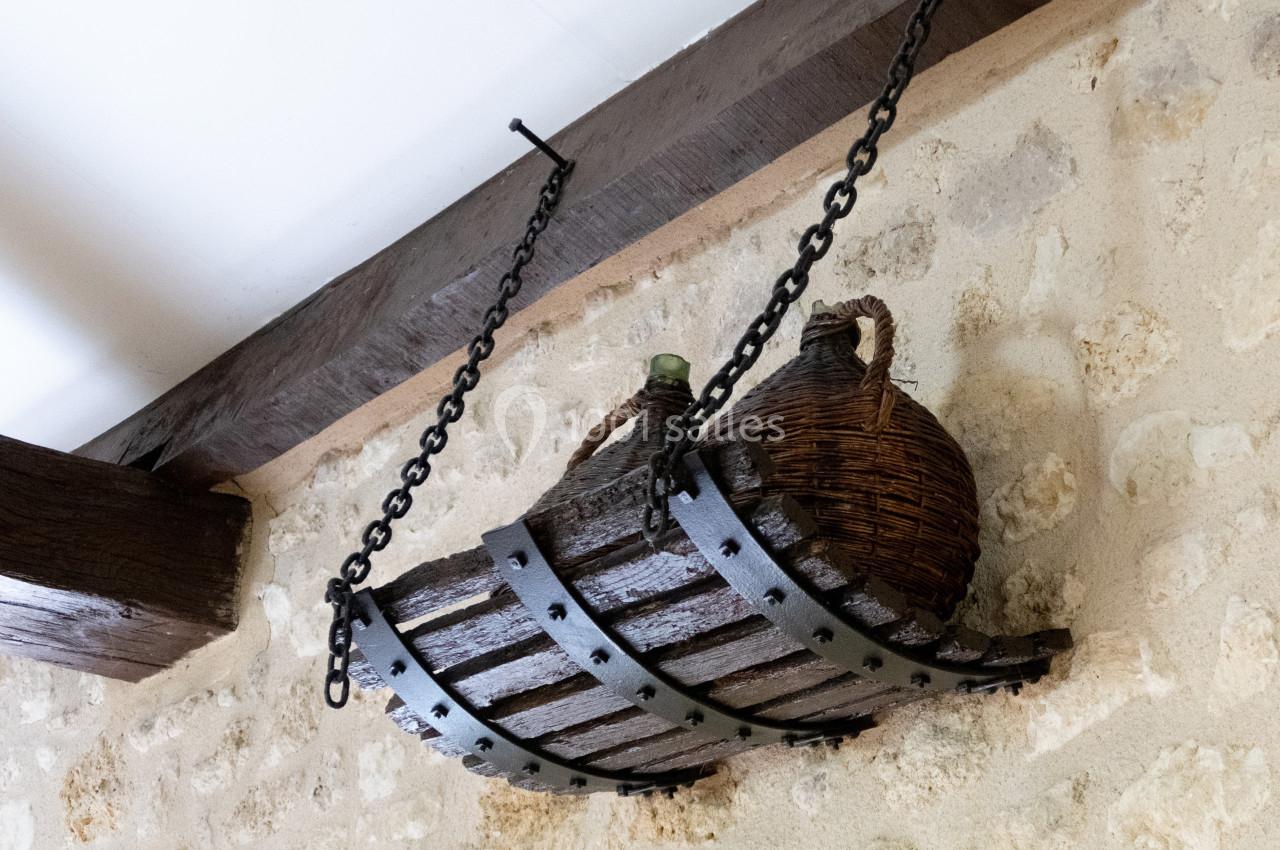 Dame-jeanne en verre protégée par un panier en osier suspendue à une chaîne fixée à une poutre en bois.