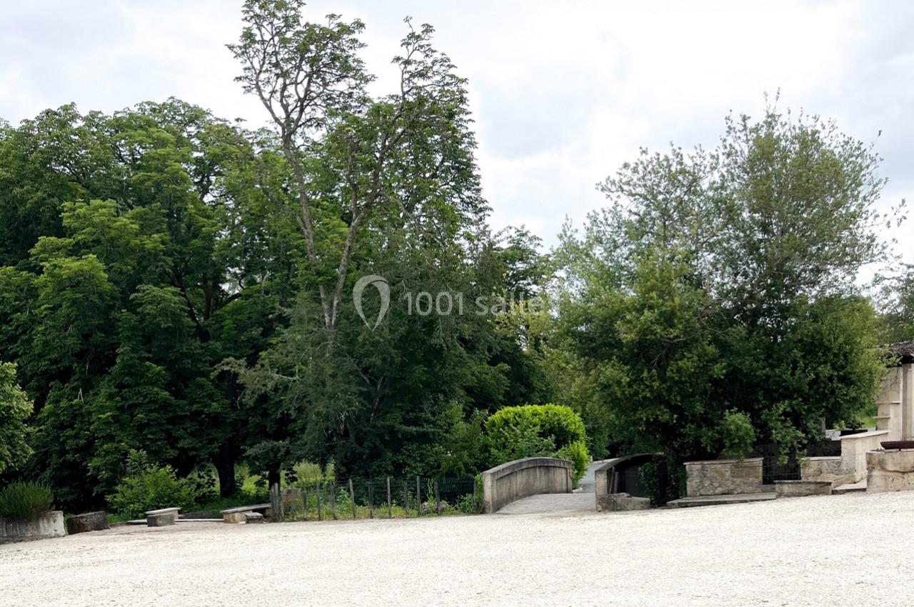 Place ouverte avec sol en gravier, entourée d'arbres verdoyants et de petits murets en pierre.