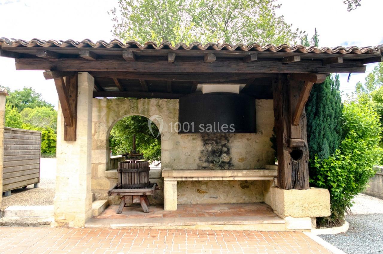Abri en pierre avec toit en tuiles, contenant un ancien pressoir en bois et un évier en pierre, entouré de végétation.