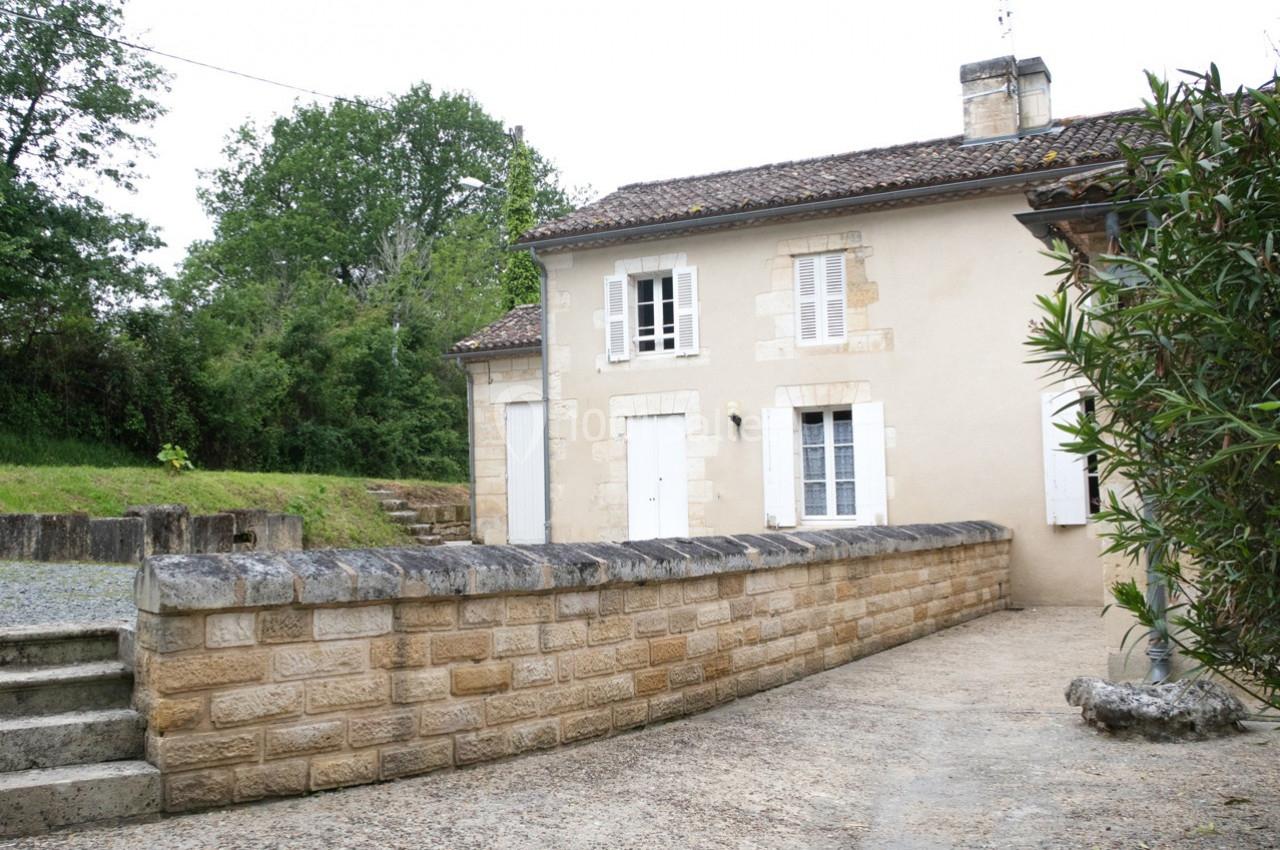 Maison en pierre beige avec volets blancs, entourée d'un muret en pierre et d'une végétation verdoyante.