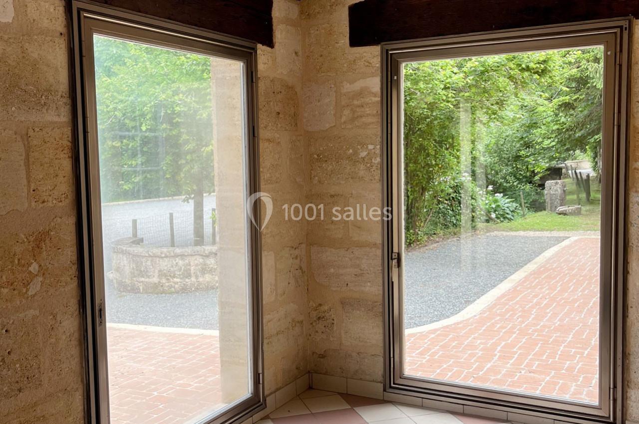 Deux grandes fenêtres donnant sur une allée pavée et un jardin verdoyant, vues depuis une pièce en pierre.